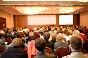 A conference room full of attendees all focused on the presenter at the front of the room.