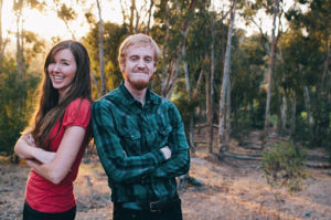 Two volunteers standing shoulder to shoulder. Both are smiling and are standing in front of a wooded area.