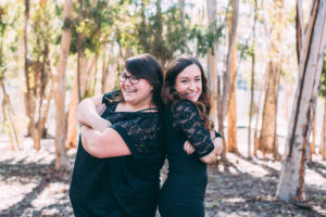 Two individuals leaning back to back, smiling at the camera in front of a wooded area.