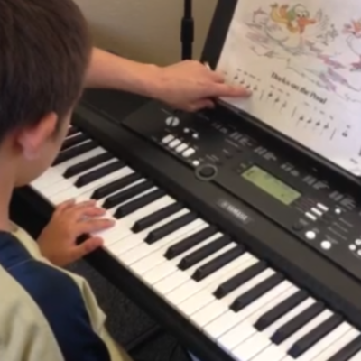 A child sits at a piano keyboard while a music therapist points to the notes on a music book.