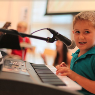 A young child sits at a piano keyboard while singing into a microphone. He looks directly at the camera.