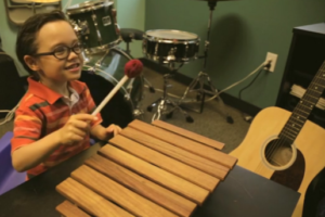 A music therapy client sitting at a xylophone, playing with a mallet and smiling at a person off camera.