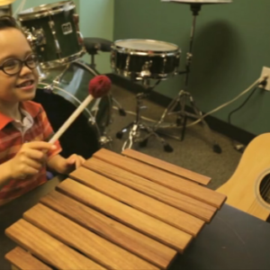 A music therapy client sitting at a xylophone, playing with a mallet and smiling at a person off camera.