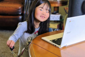 A young child sitting at a table with a laptop keyboard in front of her. She has a smile on her face and seems to be jingle a set of keys to the music on the screen.