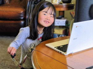 A young child sitting at a table with a laptop keyboard in front of her. She has a smile on her face and seems to be jingle a set of keys to the music on the screen.
